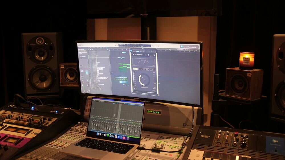 Na zdjęciu widać studio nagraniowe z dużym monitorem wyświetlającym oprogramowanie do edycji dźwięku oraz laptopem z podobnym interfejsem. W tle znajdują się głośniki i sprzęt audio, a na biurku stoi zapalona świeca.