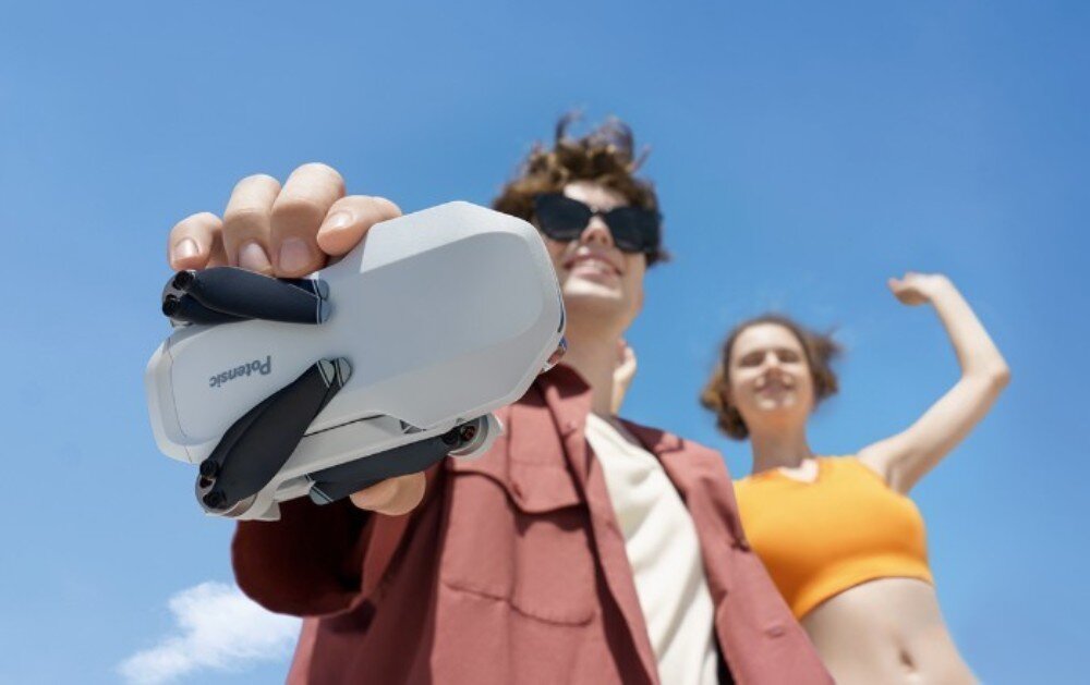 Dron POTENSIC Atom Body Dłoń trzymająca złożony mały biały dron na tle nieba i niewyraźnych postaci w oddali