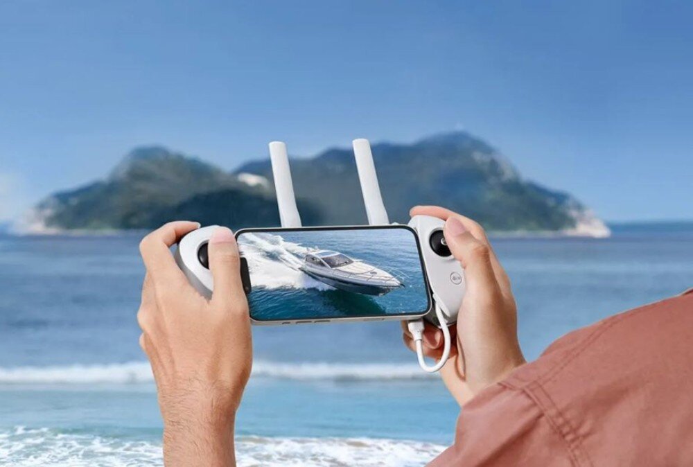 Dron POTENSIC Atom Body Dłonie trzymające biały kontroler z podłączonym telefonem wyświetlającym podgląd łodzi na morzu