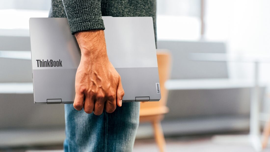 Mężczyzna w zielonej kurtce trzymający laptop Lenovo ThinkBook 14 2-in-1 w trybie zamkniętym na tle miejskiej architektury z wysokimi budynkami, podkreślający mobilność i niezawodność urządzenia w środowisku biznesowym. - Intel Core Ultra AI