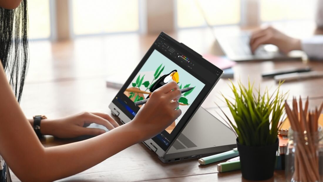 Kobieta w jasnej bluzie pracująca na laptopie Lenovo ThinkBook 14 2-in-1 w trybie tabletu z piórkiem cyfrowym, na ekranie kolorowa aplikacja do projektowania graficznego z rysunkiem ptaka i liści, na drewnianym biurku z rośliną doniczkową w tle.