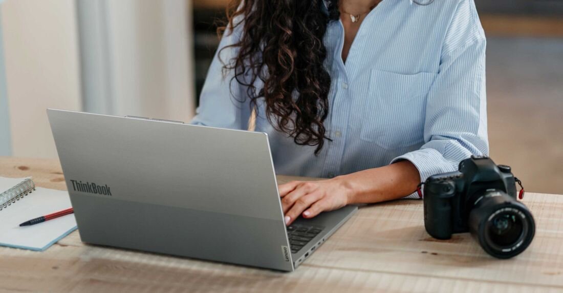 Kobieta z długimi kręconymi włosami w jasnej koszuli pracująca na laptopie Lenovo ThinkBook 16 w srebrnej obudowie z logo ThinkBook na pokrywie, obok aparat fotograficzny, na drewnianym biurku w naturalnym oświetleniu, podkreślający profesjonalne zastosowania z zabezpieczeniem TPM 2.0. - TPM 2.0 oraz czytnik linii papilarnych