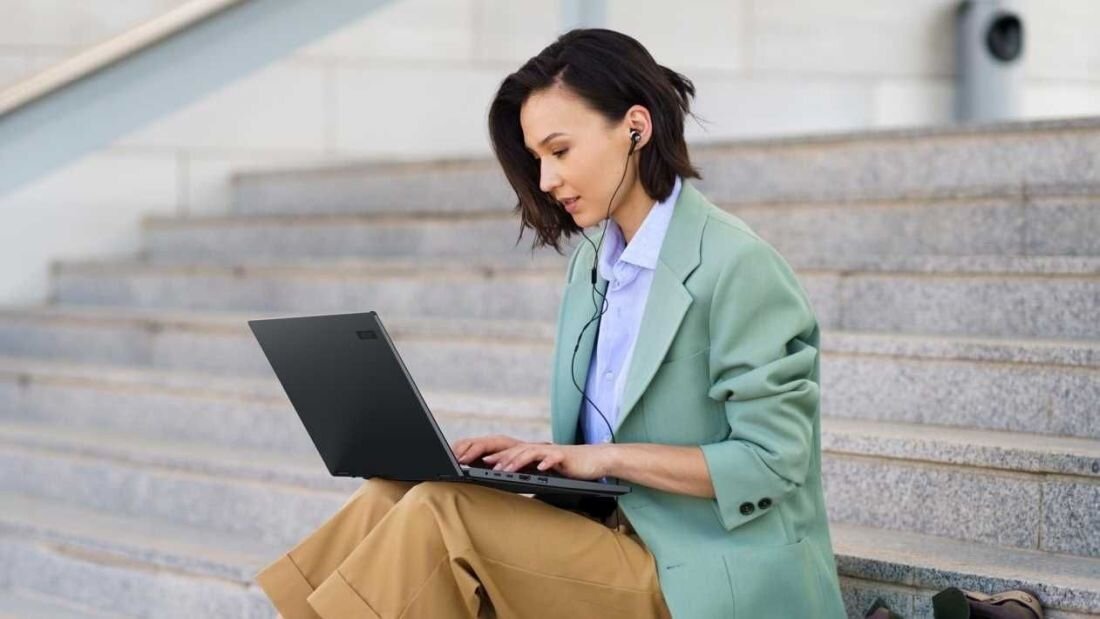 Kobieta w zielonej kurtce pracuje na laptopie ACER TravelMate P4 Spin 14, siedząc na schodach. Konstrukcja 2-w-1, Długotrwała bateria