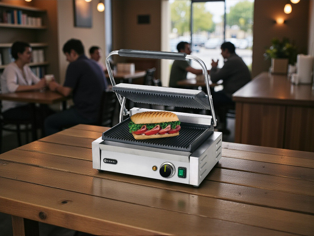 Grill elektryczny HENDI Panini 263655, kuchnia, grillowanie, gotowanie, smażenie, Grill kontaktowy Hendi 263655, otwarta pokrywa, grillowanie kanapki typu panini – urządzenie na drewnianym stole w kawiarni, w tle klienci.