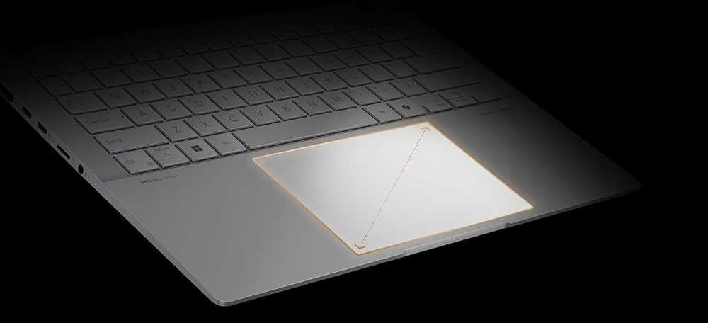 Fragment laptopa ASUS Vivobook S 16 OLED na czarnym tle, widoczny duży, prostokątny touchpad z pomarańczową ramką podświetlenia wokół krawędzi, nad nim fragment klawiatury z czarnymi klawiszami, całość podkreśla zaawansowane funkcje dotykowe urządzenia. - Rozbudowany touchpad
