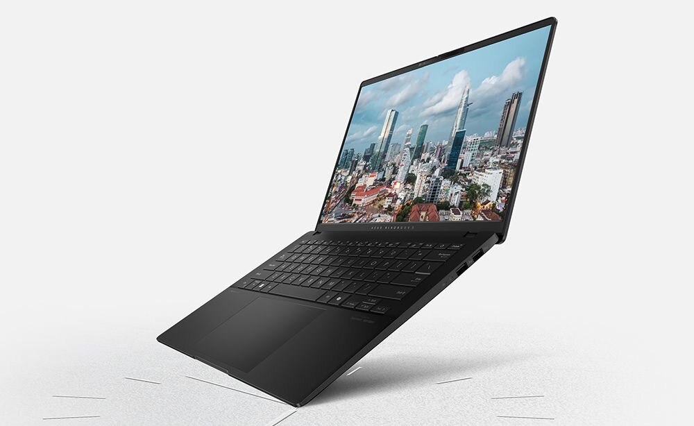 Otwarty laptop ASUS Vivobook S 16 OLED w kolorze czarnym ustawiony pod kątem na jasnym tle, na ekranie panorama miejska z wieżowcami i niebieskim niebem z białymi chmurami, cienkie ramki ekranu, minimalistyczny design obudowy. - bateria 75 Wh