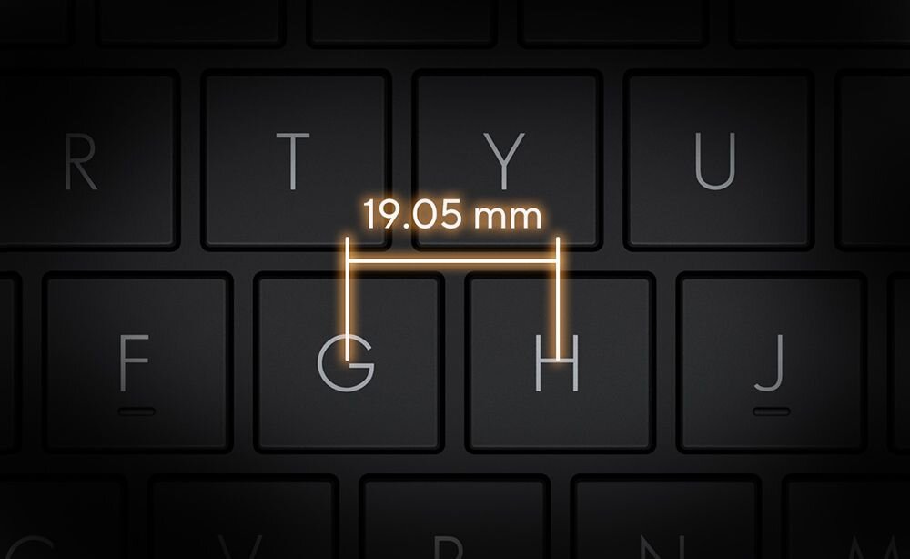 Zbliżenie na fragment czarnej klawiatury laptopa z podświetlonymi krawędziami klawiszy G i H, między nimi pomarańczowa linia z oznaczeniem 19.05 mm wskazującą standardowy odstęp między środkami klawiszy, podkreślający ergonomiczne rozmieszczenie i komfort pisania. - pełnowymiarowy rozstaw klawiszy, Długi skok klawisza