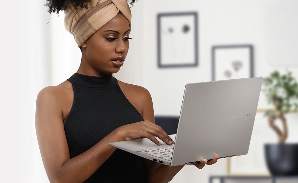 Kobieta w czarnej bluzce bez rękawów z beżowym turbanem na głowie trzyma w dłoniach srebrnego laptopa ASUS Vivobook S 16 OLED, patrzy na ekran z koncentracją, w tle jasne wnętrze z obrazami w ramkach i rośliną doniczkową, naturalne światło z okna. - Opis serii Vivobook.