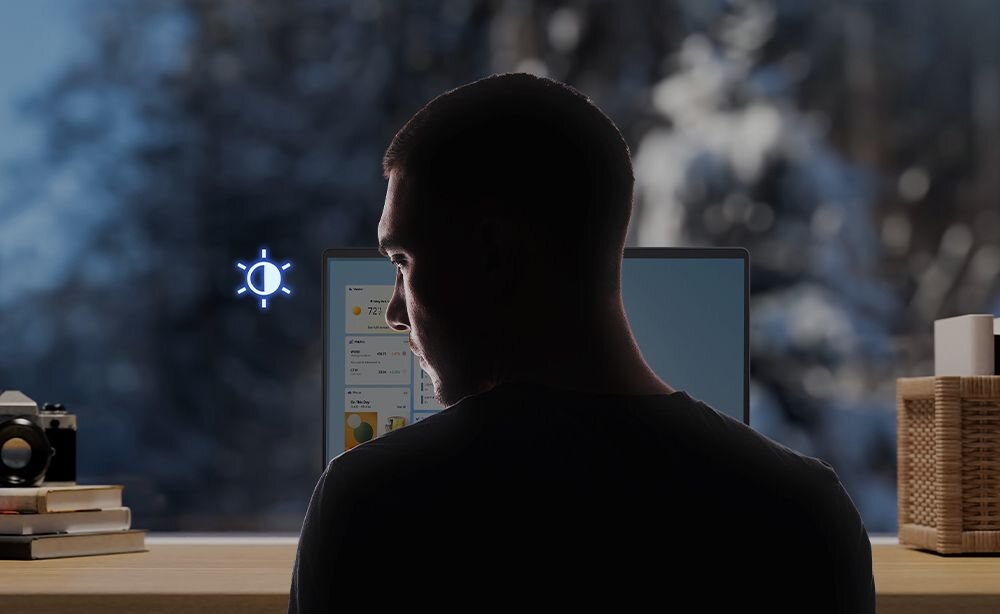 Mężczyzna w ciemnej koszuli siedzi przy biurku z laptopem ASUS Vivobook S 16 OLED, z ekranu biegną niebieskie linie skanowania w kierunku jego twarzy, symbolizujące funkcję ASUS Adaptive Dimming dostosowującą jasność ekranu do warunków oświetlenia i pozycji użytkownika, na biurku książki, aparat fotograficzny i drewniane akcesoria. - ASUS Adaptive Dimming