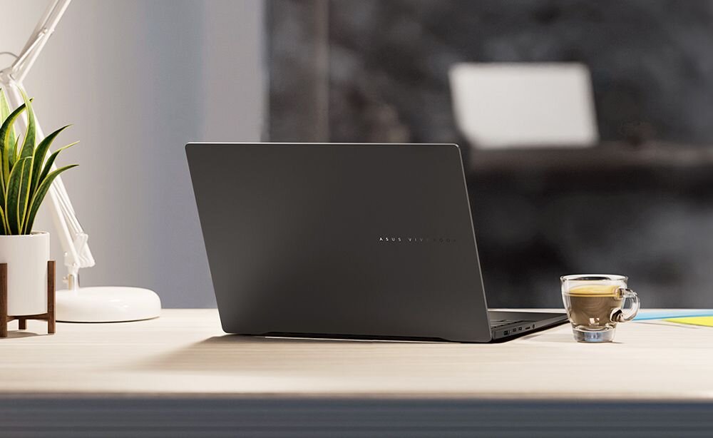 Zamknięty laptop ASUS Vivobook S 16 OLED w kolorze grafitowym ustawiony na jasnym, drewnianym biurku w nowoczesnym wnętrzu; obok przezroczysta szklanka z kawą latte, po lewej biała doniczka z zieloną rośliną sansewieria, w tle rozmyte wnętrze z białymi meblami i szarą ścianą, naturalne światło z okna. - eleganckie wzornictwo