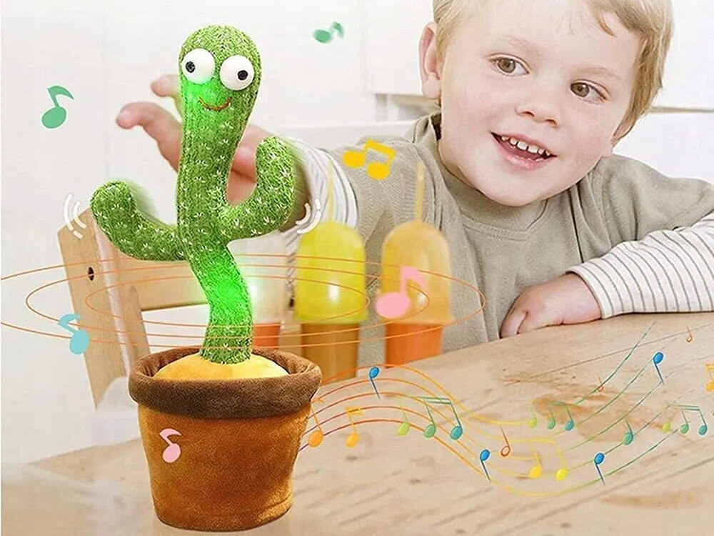 Zabawka interaktywna EXTRALINK Kaktus EX.24244 Uśmiechnięte dziecko obserwujące interaktywną zabawkę tańczący kaktus, która świeci się i porusza na stole w otoczeniu kolorowych grafik nutek.