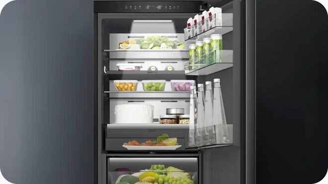Otwarta lodówka z widocznymi półkami wypełnionymi różnymi produktami spożywczymi, takimi jak warzywa, napoje i słoiki. Wnętrze jest dobrze oświetlone, a drzwi lodówki mają dodatkowe półki na butelki i kartony.