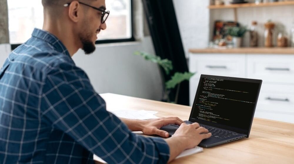 Mężczyzna w okularach programujący na laptopie ASUS ExpertBook B5 przy drewnianym biurku, w tle nowoczesne biuro, podkreślający profesjonalne zastosowanie i bezpieczeństwo dzięki czytnikowi linii papilarnych . - czytnik linii papilarnych