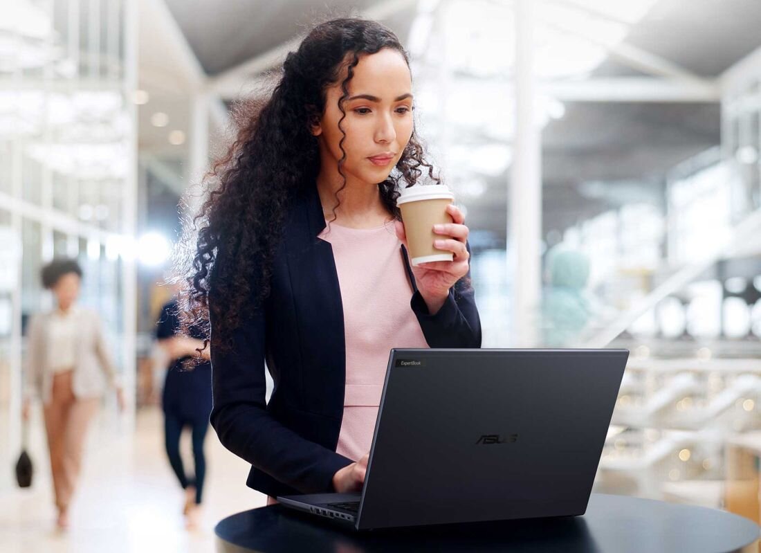 Kobieta z kręconymi włosami pracująca na laptopie ASUS ExpertBook B5, trzymając kubek z kawą w lobby biurowym, podkreślający płynną łączność dzięki Wi-Fi 6 dla mobilnych profesjonalistów . - Wi-Fi 7
