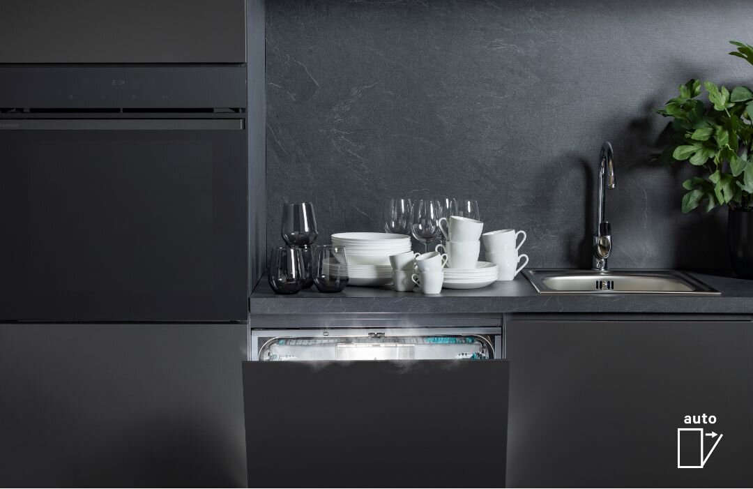 Kuchnia z otwartą zmywarką, na blacie stoją naczynia i szklanki. Obok zlewozmywak i roślina, w rogu widoczny symbol z napisem \'auto\'.
