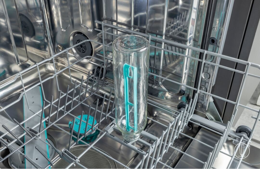 Wnętrze zmywarki z umieszczoną w środku szklaną butelką z turkusowym uchwytem. W tle widoczne są metalowe elementy konstrukcji zmywarki.