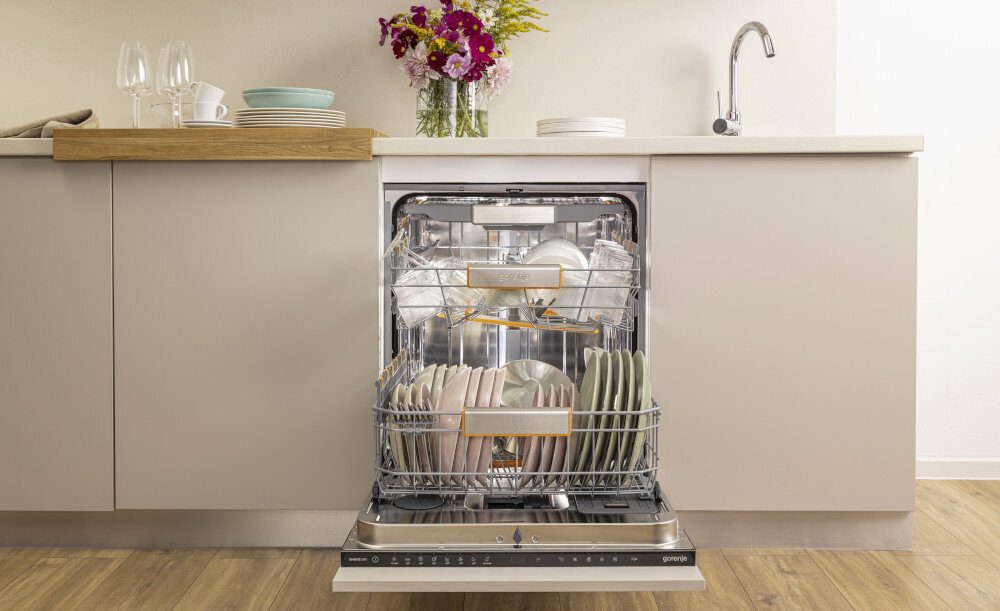 ZMYWARKA GORENJE GV693A65AD Na zdjęciu jest otwarta zmywarka w zabudowie kuchennej, wypełniona czystymi naczyniami. Na blacie stoi zestaw naczyń, szklanek oraz wazon z kolorowymi kwiatami
