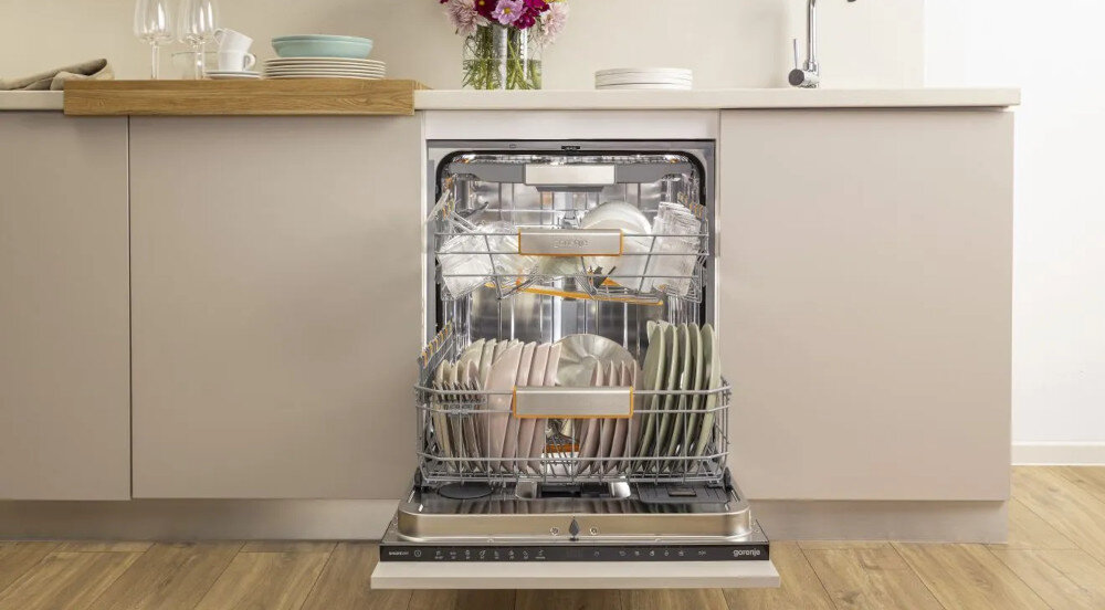 ZMYWARKA GORENJE GV693A65AD Na zdjęciu znajduje się otwarta zmywarka w zabudowie kuchennej, wypełniona czystymi naczyniami różnego typu. W tle widoczny jest jasny blat z zastawą i bukietem kwiatów ustawionym przy zlewozmywaku zmywarka elastyczne wnętrze trzy kosze 16 nakryć duży załadunek imprezy