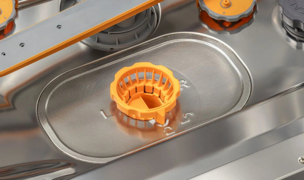 ZMYWARKA GORENJE GV693A65AD Na zdjęciu pokazano dolną część wnętrza zmywarki, gdzie znajduje się metalowy filtr z pomarańczowym uchwytem. W tle widoczne są ramiona spryskujące oraz elementy techniczne zmywarki mniej czyszczenia