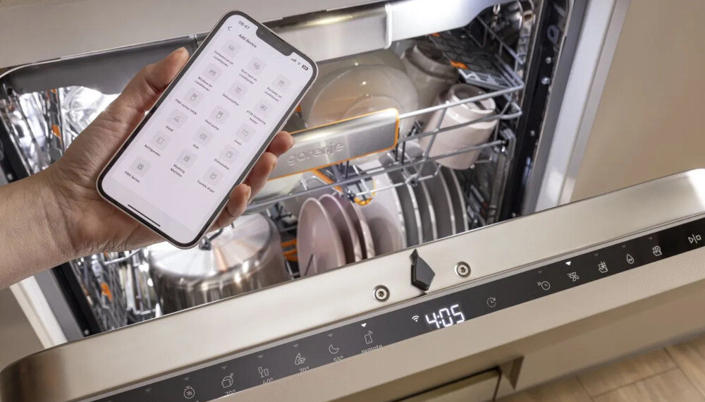 ZMYWARKA GORENJE GV693A65AD Zdjęcie przedstawia dłoń trzymającą smartfon z aplikacją do zdalnego sterowania zmywarką, a w tle otwarta zmywarka z czystymi garnkami i talerzami w środku. Panel sterowania zmywarki pokazuje czas pracy, a widoczna aplikacja podkreśla wygodę obsługi urządzenia zdalne sterowanie ConnectLife powiadomienia funkcje mobilne