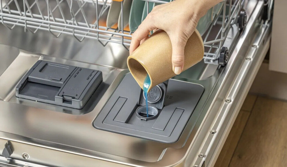 ZMYWARKA GORENJE GV693A65AD Na zdjęciu widać otwartą zmywarkę, do której ręka nalewa płyn nabłyszczający do specjalnego dozownika. W tle widoczne są czyste naczynia ustawione w koszach urządzenia SmartDosing zmywarka oszczędność czas detergent energia automatyczne dozowanie pojemnik łatwe czyszczenie 20 cykli