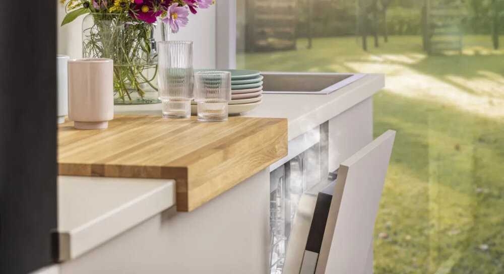 ZMYWARKA GORENJE GV693A65AD Na zdjęciu widać kuchenny blat z bukietem kwiatów, szklankami i talerzami ustawionymi obok zlewu. Na pierwszym planie znajduje się otwarta zmywarka, a w tle przez duże okno widać ogród zmywarka szybkie suszenie niskie zużycie energii automatyczne otwieranie drzwi świeże powietrze suche naczynia plastik