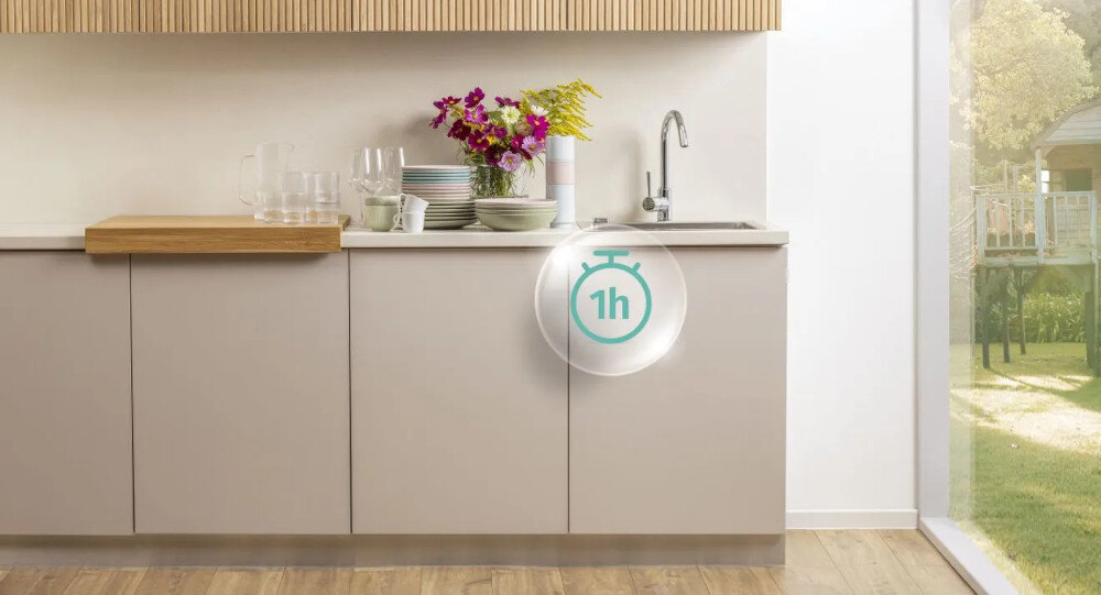 ZMYWARKA GORENJE GV693A65AD Na zdjęciu znajduje się jasna kuchnia z bukietem kwiatów i zastawą na blacie, z widocznym zlewem pod ścianą. Po prawej stronie kuchni widać duże okno z widokiem na ogród, a na frontach szafek umieszczona jest grafika z ikoną zegara „1h”, sugerującą szybki program zmywarki zmywarka szybki program codzienne mycie 65°C 1 godzina oszczędność czasu lekkie zabrudzenia
