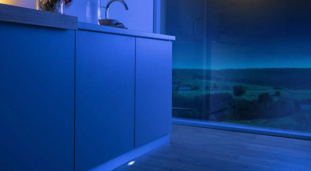 ZMYWARKA GORENJE GV693A65AD Na zdjęciu widać kuchnię oświetloną niebieskim światłem, z dużym oknem wychodzącym na nocny krajobraz. Na blacie ustawione są naczynia i wazon z kwiatami, a na pierwszym planie widoczne są zamknięte szafki kuchenne zmywarka wskaźnik LED faza cyklu informacja praca zmywarki koniec cyklu