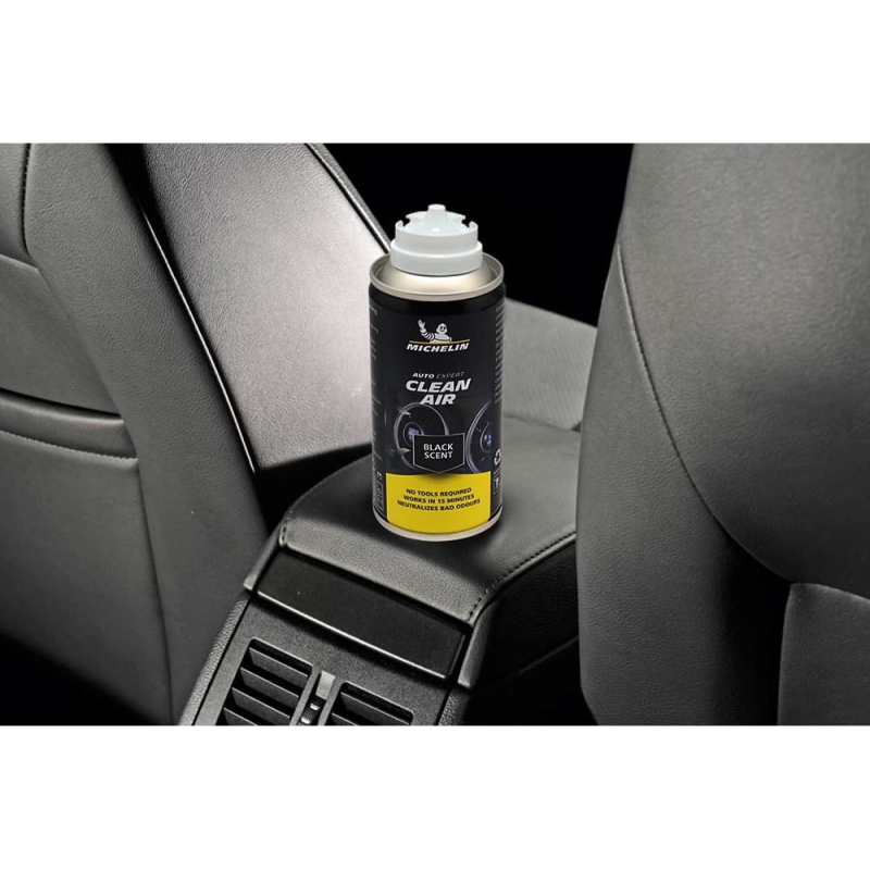Na zdjęciu znajduje się puszka produktu \'Michelin Auto Expert Clean Air Black Scent\' umieszczona na konsoli środkowej samochodu. Puszka ma czarną etykietę z białym i żółtym tekstem, a w tle widoczne są elementy wnętrza samochodu.