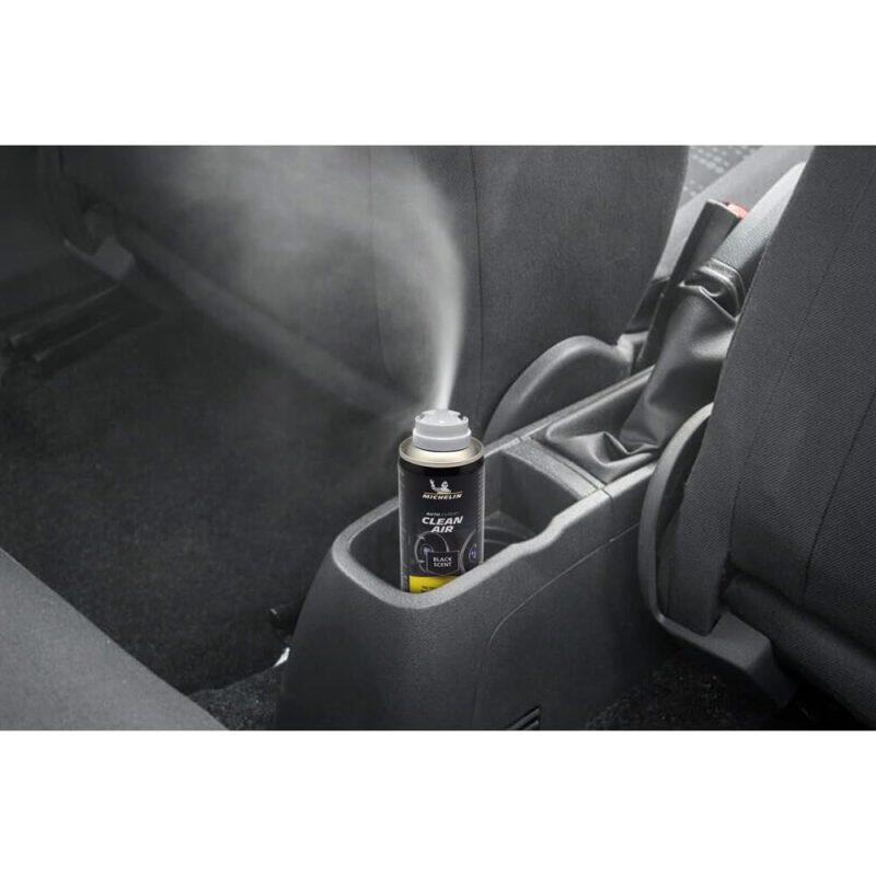 W samochodowym uchwycie na kubki znajduje się puszka z napisem \'MICHELIN CLEAN AIR BLACK SCENT\', z której wydobywa się mgiełka. W tle widoczne są siedzenia samochodowe.