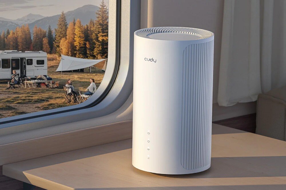 Router CUDY P4 5G Widok routera stojącego na blacie przy oknie w kamperze. Za oknem widać grupę osób siedzących na zewnątrz obok przyczepy kempingowej, w tle jesienny las i góry, opis