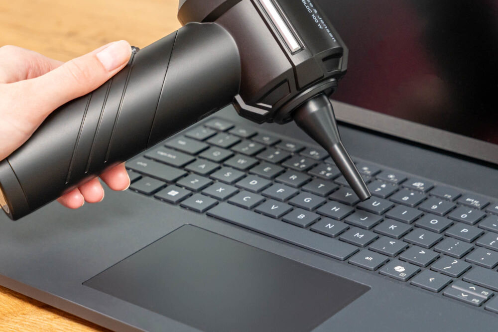 Odkurzacz ręczny GÖTZE & JENSEN HV400 Odkurzacz ręczny z wąską dyszą ssącą używany do czyszczenia klawiatury laptopa. Dłonie trzymają urządzenie nad czarnymi klawiszami.