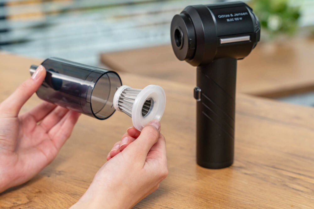 Odkurzacz ręczny GÖTZE & JENSEN HV400 Dłonie wyjmują metalowy filtr siatkowy z przezroczystego pojemnika na kurz odkurzacza, demonstrując proces czyszczenia lub konserwacji.