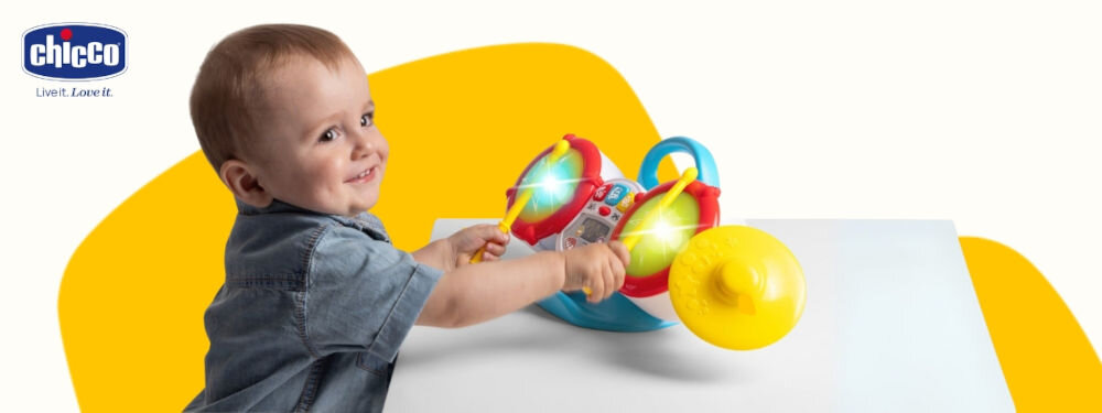 Zabawka perkusja CHICCO 00012243000000 biało żółte tło maluch trzyma pałeczki uderza w perkusję kolorowe elementy przyciski świecące bębenki nauka przez zabawę 