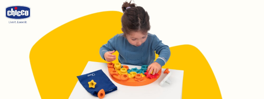 Zabawka edukacyjna CHICCO Smart2Play Kolorowe Pinezki Tęcza 00012198000000 nauka przez zabawę biało żółte tło dziecko bawi się kolorową zabawką na stoliku różne kolory i kształty obok leży ciemny woreczek