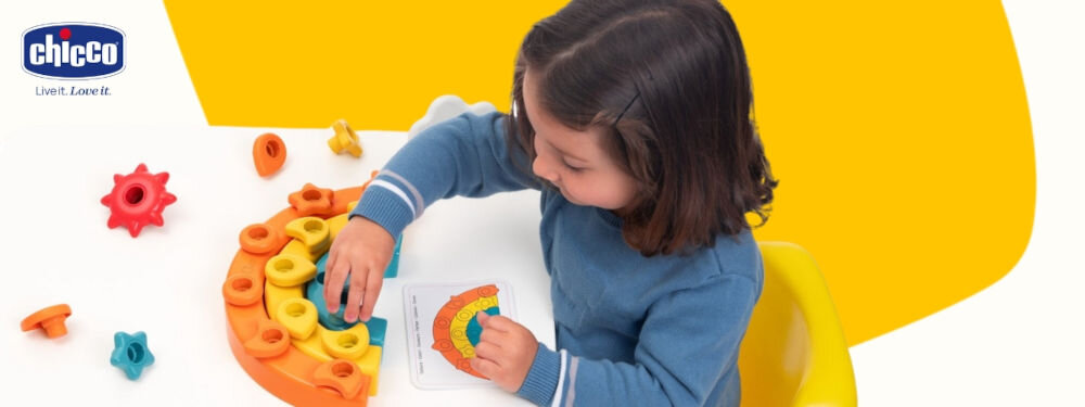 Zabawka edukacyjna CHICCO Smart2Play Kolorowe Pinezki Tęcza 00012198000000 biało żółte tło dziecko układa kolorową tęczę i patrzy na kartę z ilustracją budowli Manipulowanie elementami rozwijanie zdolności manualnych nauka kolorów, liczb i kształtów pobudzenie rozwoju poznawczego istotny element edukacji wizualnej wykonywanie zadań na kartach edukacyjnych ćwiczenie pamięci wzrokowej