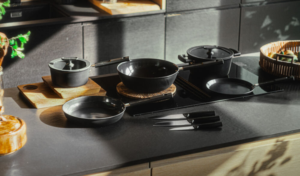 Patelnia wok FISKARS Hard Face 1075523 28 cm do każdej kuchni czarne garnki i patelnie stoją na płycie obok leżą noże