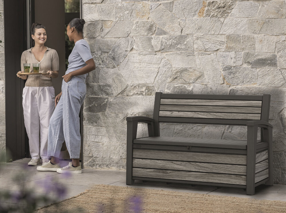 Ławka ogrodowa KETER Deco Garden Bench Brązowy ławka stoi na tle kamiennej ściany obok drzwi dwie osoby rozmawiają ze sobą jedna trzyma tacę z napojami funkcjonalna wygodna komfortowy odpoczynek szerokie siedzisko o wymiarach 126 x 52 cm miejsce do siedzenia dla dwóch osób ergonomiczne podłokietniki stabilna konstrukcja o maksymalnym udźwigu do 272 kg