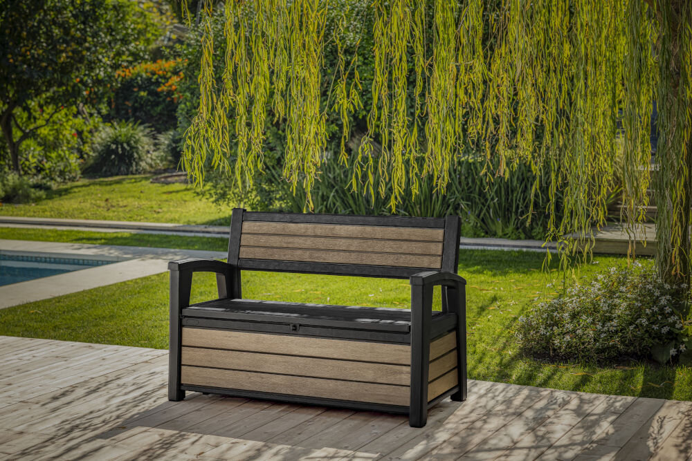 Na obrazie znajduje się ławka ogrodowa w kolorze popielatym z drewnianymi elementami, ustawiona na tle zielonych roślin. Obok ławki stoi donica z kwiatami.