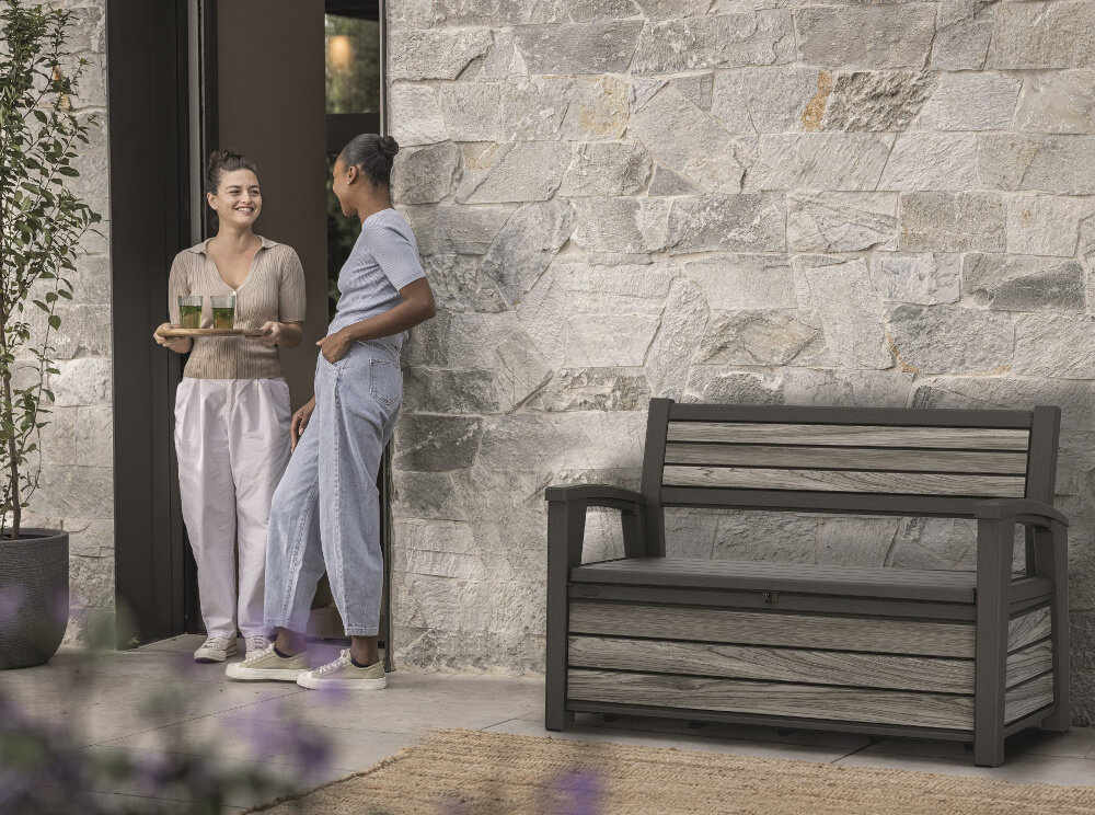 Ławka ogrodowa KETER Deco Garden Bench Szary ławka stoi na tle kamiennej ściany obok drzwi dwie osoby rozmawiają ze sobą jedna trzyma tacę z napojami funkcjonalna wygodna komfortowy odpoczynek szerokie siedzisko o wymiarach 126 x 52 cm miejsce do siedzenia dla dwóch osób ergonomiczne podłokietniki stabilna konstrukcja o maksymalnym udźwigu do 272 kg