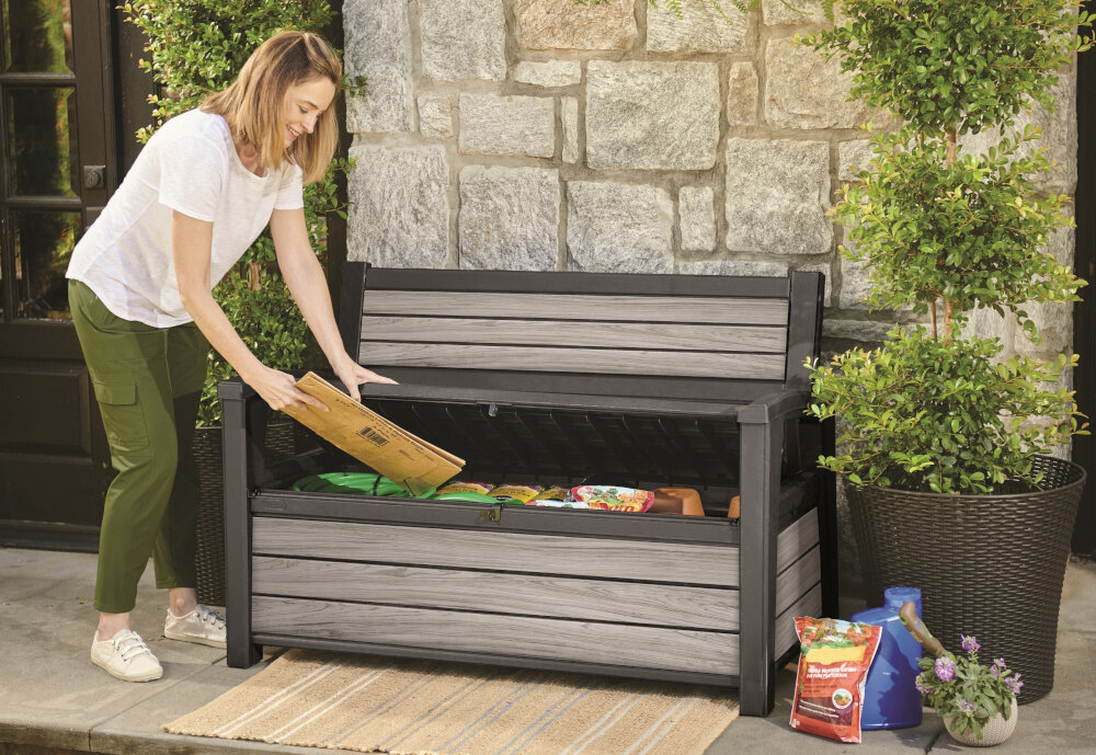 Ławka ogrodowa KETER Deco Garden Bench Szary ławka z otwartą pokrywą w środku różne rzczy osoba trzyma w dłoni złożony karton praktyczny schowek o pojemności do 227 litrów wygodne przechowywanie porządek w otoczeniu łatwe otwieranie i zamykanie zabezpieczenie na kłódkę ochrona bezpieczne przechowywanie rzecz