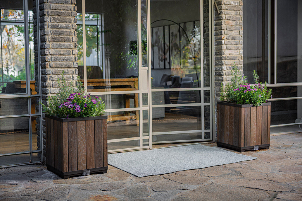 Na obrazie widoczne są dwie kwadratowe doniczki z drewnianym wykończeniem, ustawione przed przeszklonymi drzwiami. W doniczkach rosną zielone rośliny z fioletowymi kwiatami.
