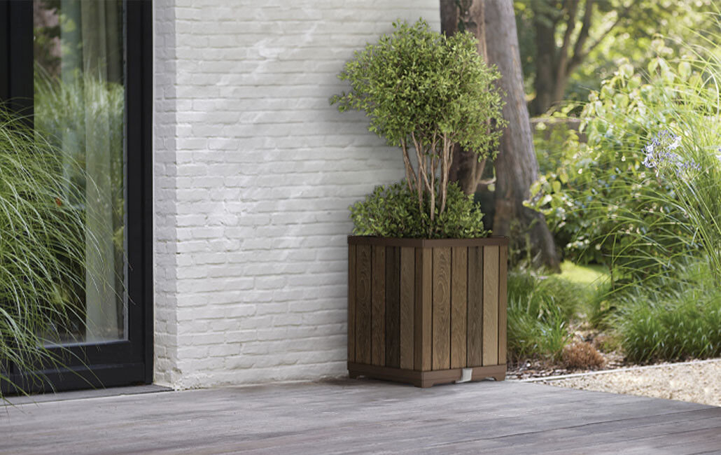 Na obrazie widoczna jest doniczka z drewnianym wzorem, w której rośnie zielony krzew. Doniczka stoi na tarasie przy białej ceglanej ścianie, otoczona zielenią ogrodu.