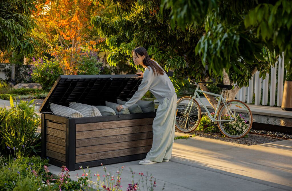 Skrzynia ogrodowa KETER Deco deck box 630 L Jasny-orzech, siłowniki, mechanizm, wygoda, zawiasy, Kobieta wkładająca poduszki do otwartej skrzyni ogrodowej Deco 630L.