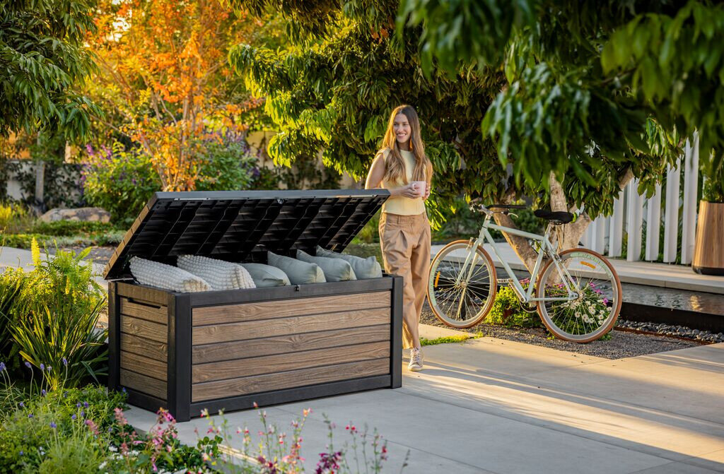Skrzynia ogrodowa KETER Deco deck box 630 L Jasny-orzech, polipropylen, wentylacja, trwałość, budowa, Kobieta przy otwartej skrzyni ogrodowej Deco wypełnionej poduszkami.