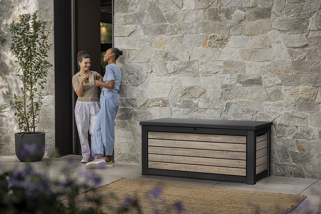 Skrzynia ogrodowa KETER Deco Deck Box 630L Brązowo-czarny, tworzywo, UV, deszcz, trwałość, Dwie kobiety rozmawiające obok skrzyni ogrodowej Deco na patio.