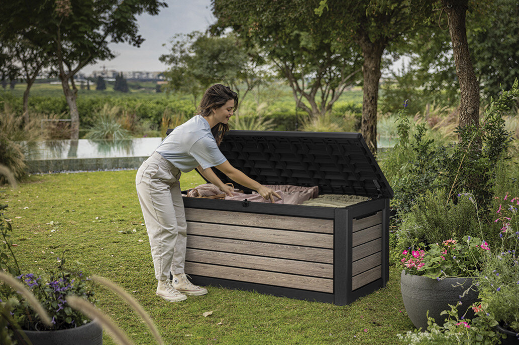 Skrzynia ogrodowa KETER Deco Deck Box 630L Brązowo-czarny, siłowniki, mechanizm, wygoda, zawiasy, Kobieta wkładająca poduszki do otwartej skrzyni ogrodowej Deco 630L.
