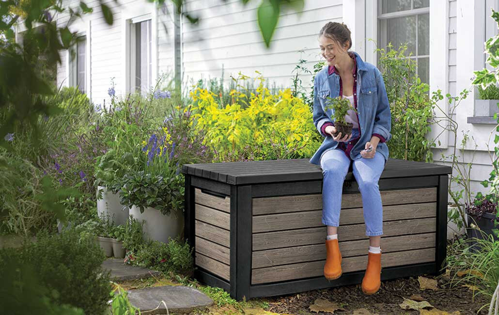 Skrzynia ogrodowa KETER Deco Deck Box 630L Brązowo-czarny, siedzenie, 272 kg, ławka, stabilność, Kobieta siedząca na skrzyni ogrodowej Keter z rośliną w doniczce.