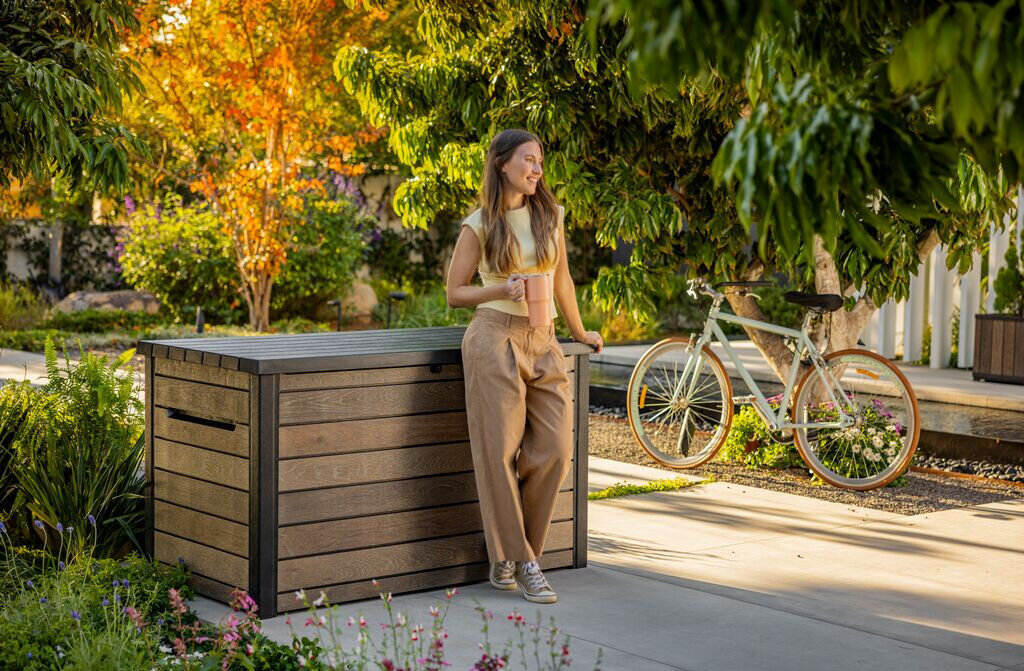 Na zdjęciu znajduje się brązowa skrzynia ogrodowa z poziomymi panelami, obok której stoi osoba trzymająca kubek. W tle widoczny jest rower oraz drzewa i rośliny w ogrodzie.