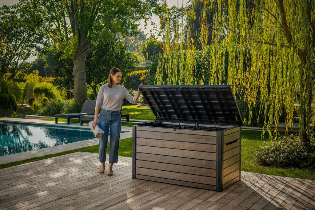 Kobieta stoi na drewnianym tarasie obok otwartej, brązowej skrzyni ogrodowej. W tle widoczny jest basen i zielony ogród z drzewami.